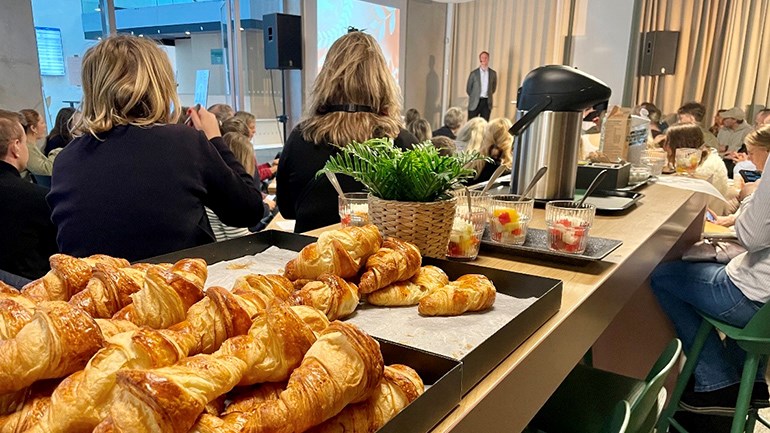 Personer som lyssnar på föreläsning med fika i förgrunden.