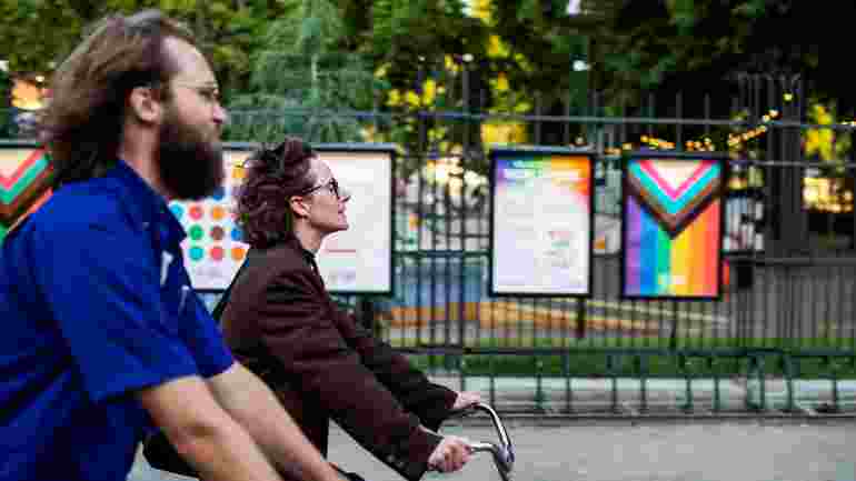 Två personer cyklar fram på gata utanför Folkets park. I Bakgrunden syns afficher, bland annat med regnbågs- och transflaggan. 