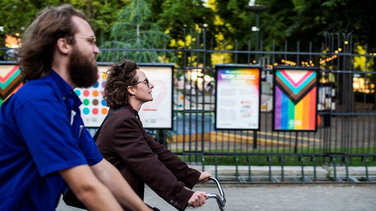 Två personer cyklar fram på gata utanför Folkets park. I Bakgrunden syns afficher, bland annat med regnbågs- och transflaggan.