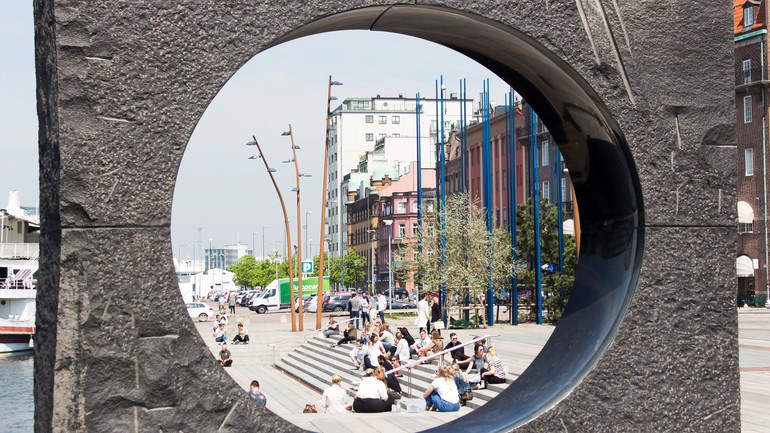 Människor sitter i solen på posthusplatsen. I förgunden syns konstverket Utblick / Insikt.